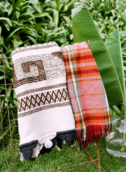 Two woven blankets hanging on a stand outdoors, one white and brown, one orange plaid.