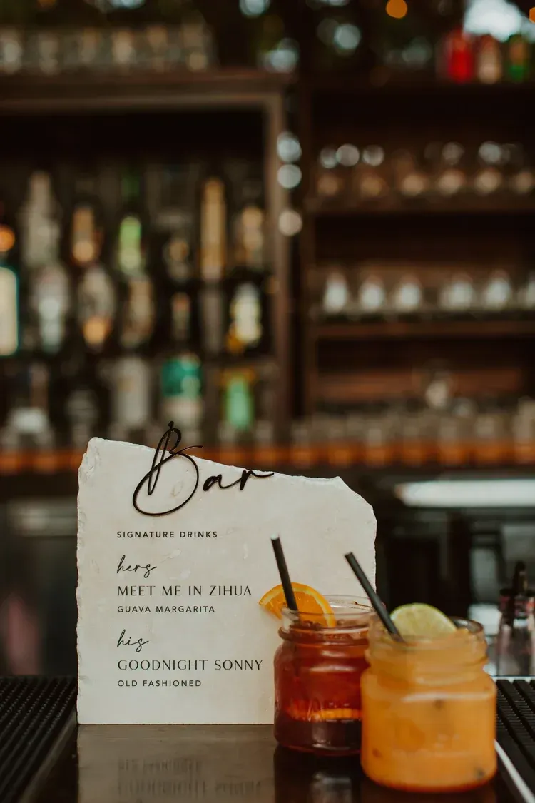 Cocktail menu card beside two orange cocktails on a bar counter, with blurred bottles in the background