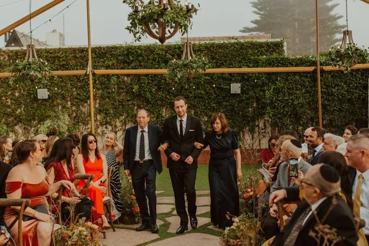 Wedding party walking down an outdoor aisle under greenery, guests seated on both sides.