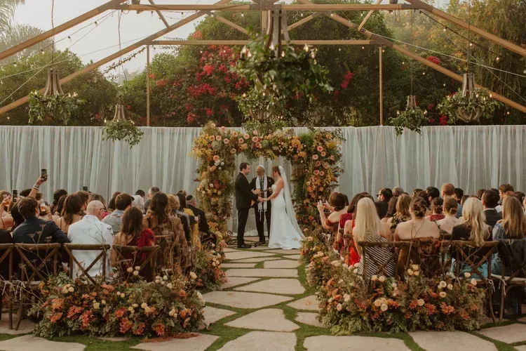 Outdoor wedding ceremony under a floral arch in a greenhouse, with guests seated on both sides.