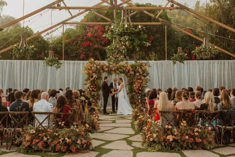 Wedding ceremony under a floral arch in a greenhouse, with guests seated on both sides.