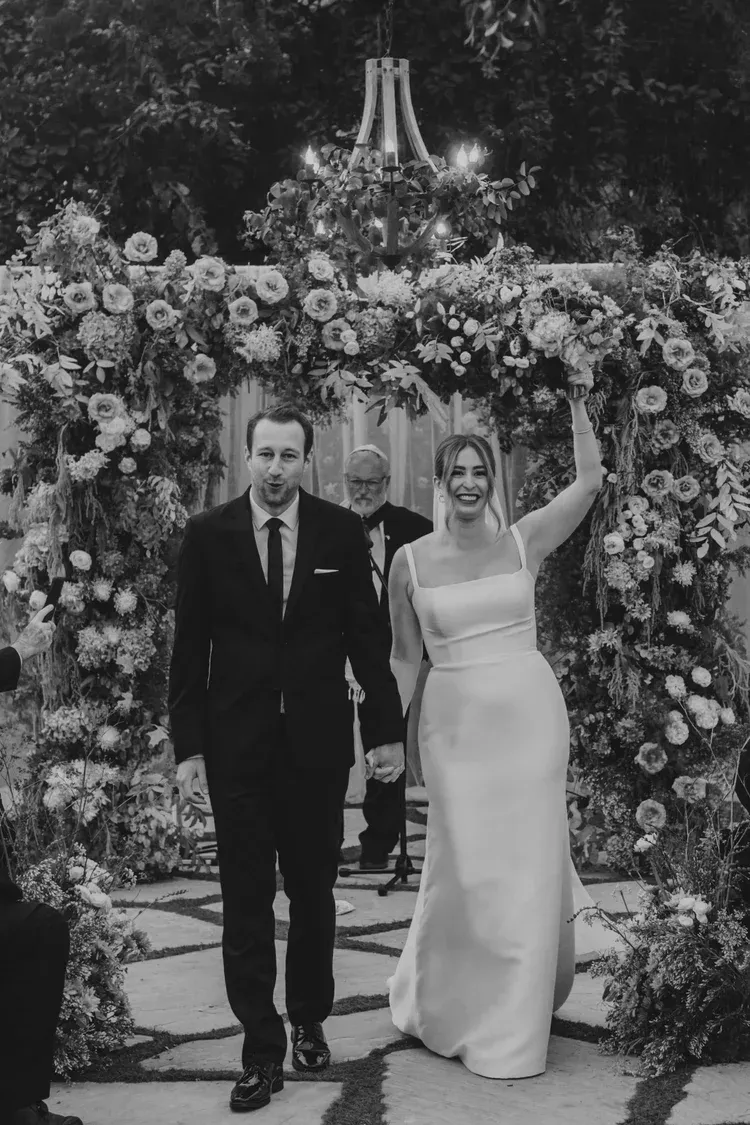 Bride and groom walking under a flower-covered arch, smiling and waving at their wedding.
