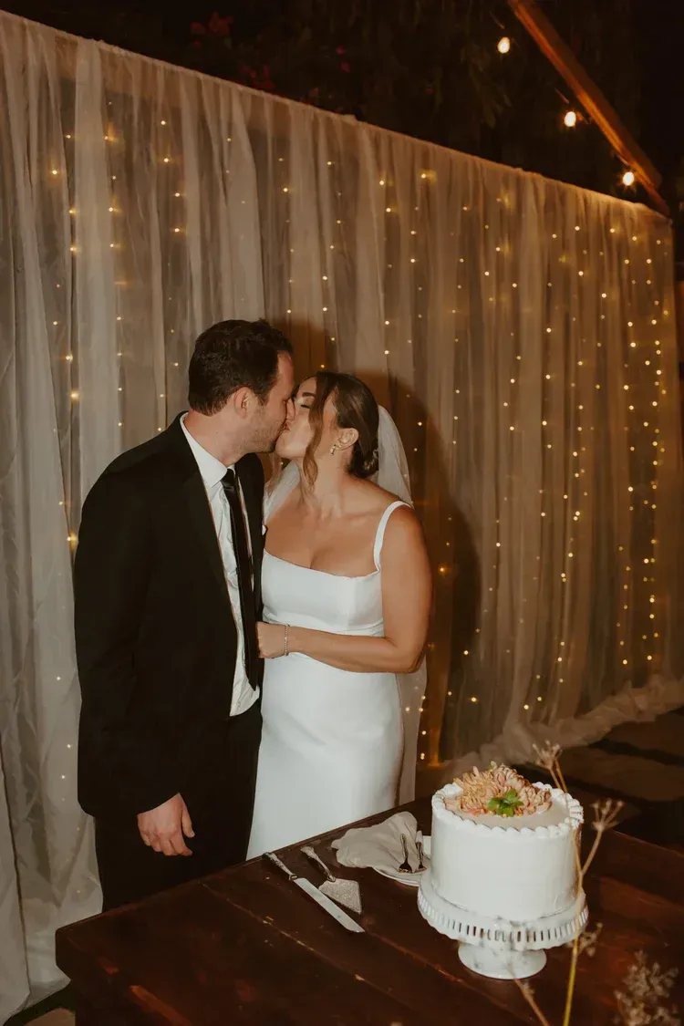 Newlyweds kissing beside a wedding cake, with white drapes and warm string lights in the background