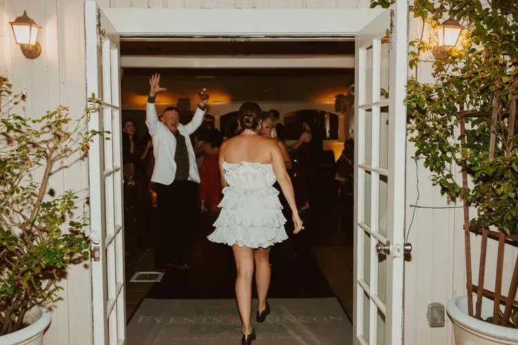 Woman in a white dress entering a dimly lit party venue through open doors while guests dance inside