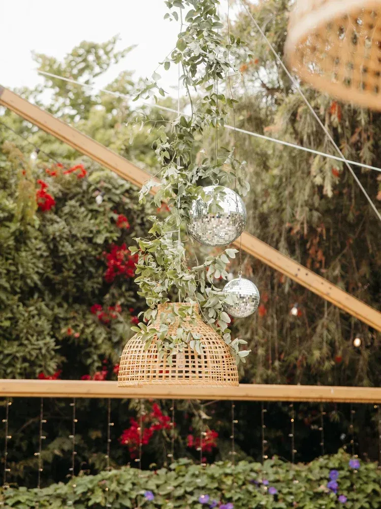 Outdoor garden setup with hanging glass ornaments, woven lantern, and string lights among greenery and flowers