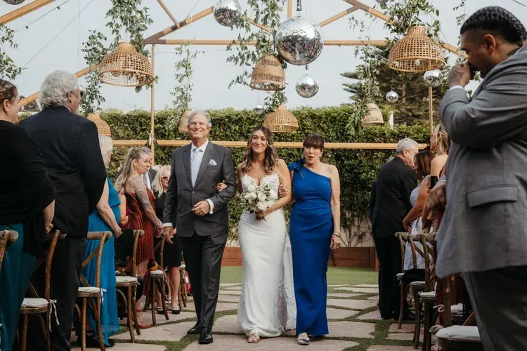 Wedding ceremony with bride and escort walking down an outdoor aisle under hanging lanterns
