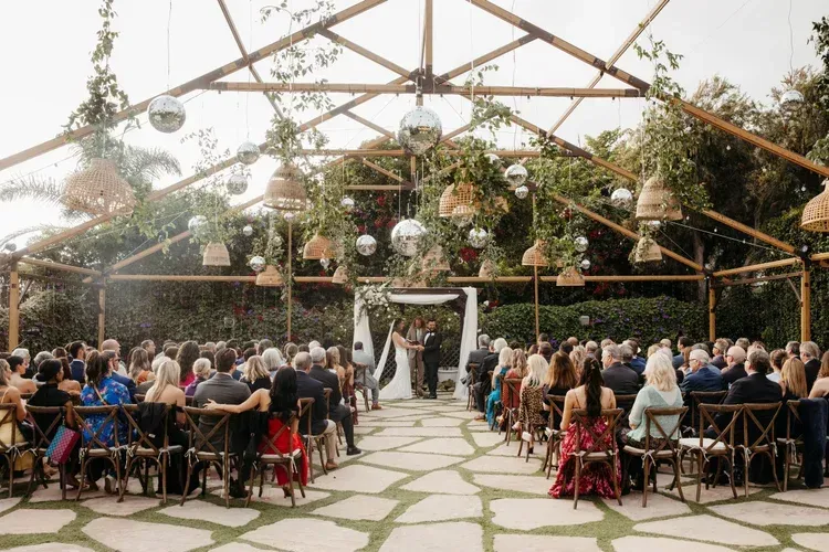 Outdoor wedding ceremony under a decorated greenhouse roof with seated guests and aisle flowers