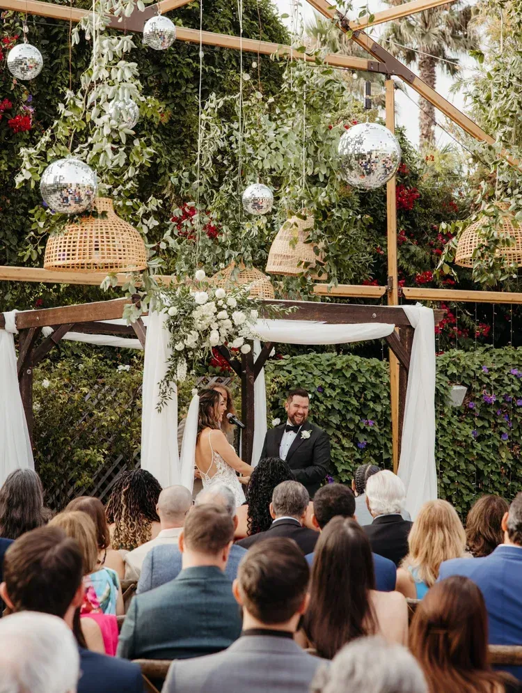 Outdoor wedding ceremony under a floral arch with guests seated and an officiant at the altar.