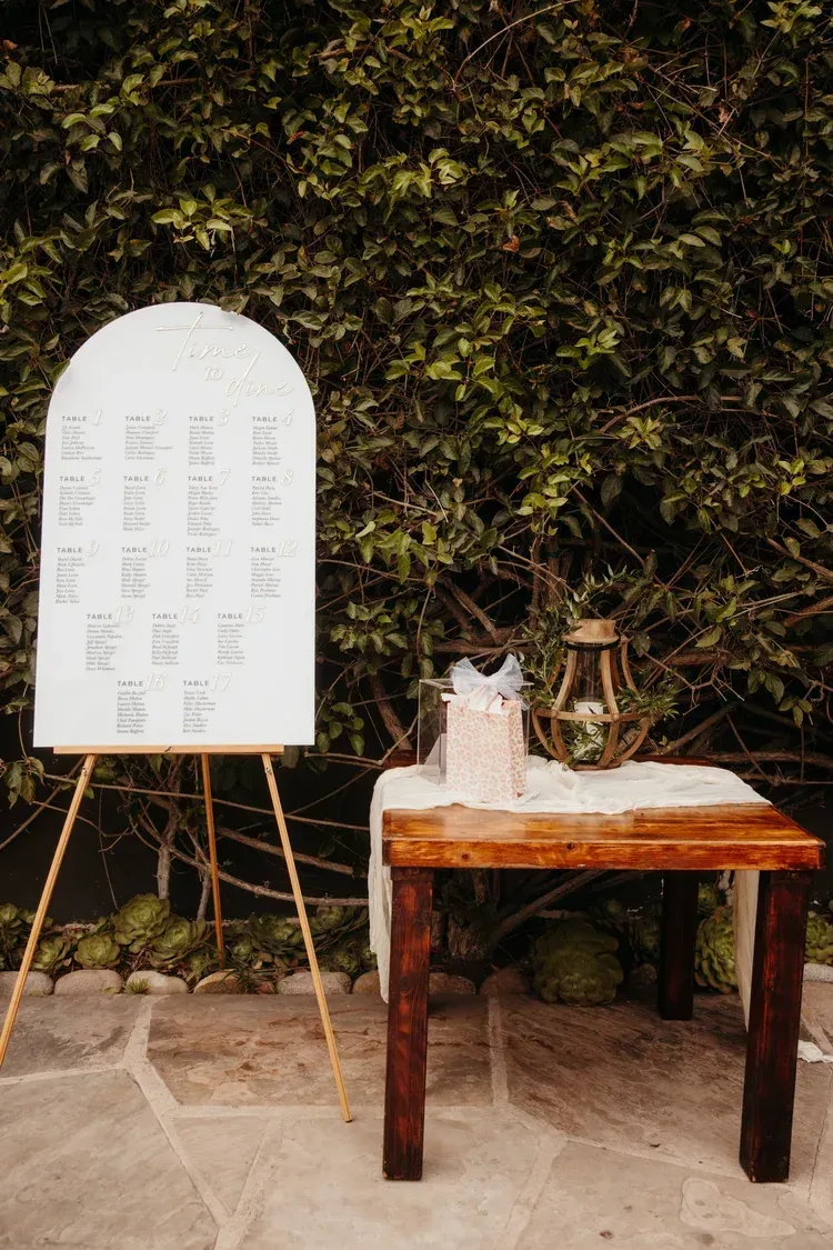 Small table with a white cloth and items beside a white sign on an easel in a garden courtyard.