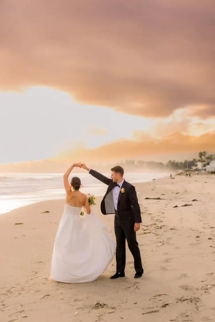 Couple dancing on a beach at sunset, with waves, palm trees, and a glowing sky.
