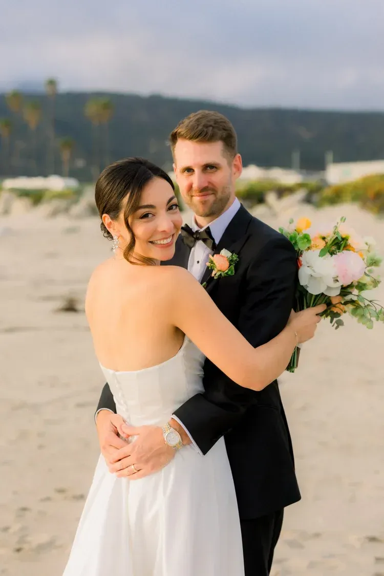 Newlyweds embracing on a sandy beach, with a bouquet and soft mountains in the background