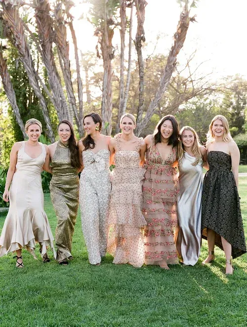 Six women in formal dresses standing arm-in-arm on a grassy lawn at sunset.
