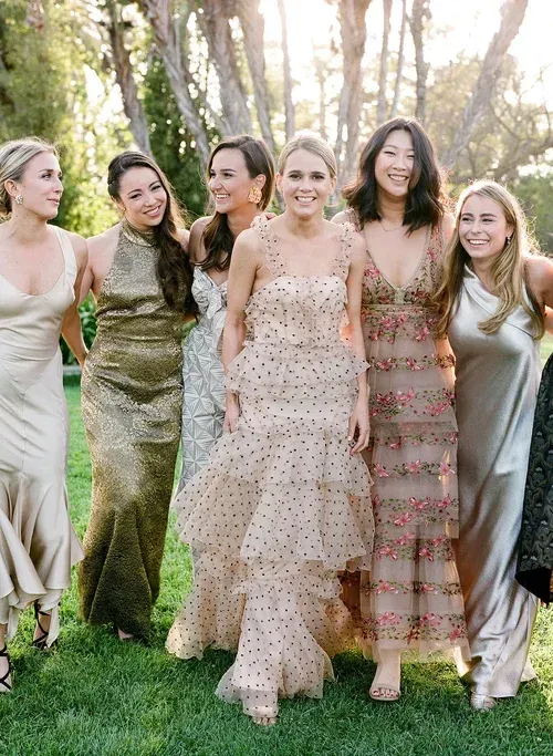 Six women in pastel dresses standing together outdoors in a sunlit garden