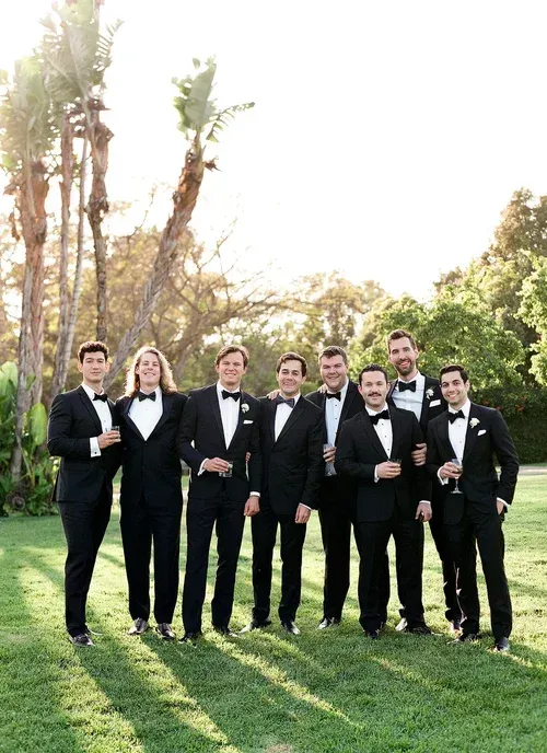 Seven people in black formalwear standing on grass in a sunny garden, posing for a group photo