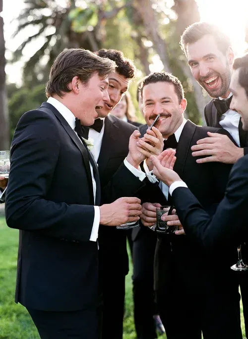 Four men in suits smiling and toasting drinks outdoors in sunlight