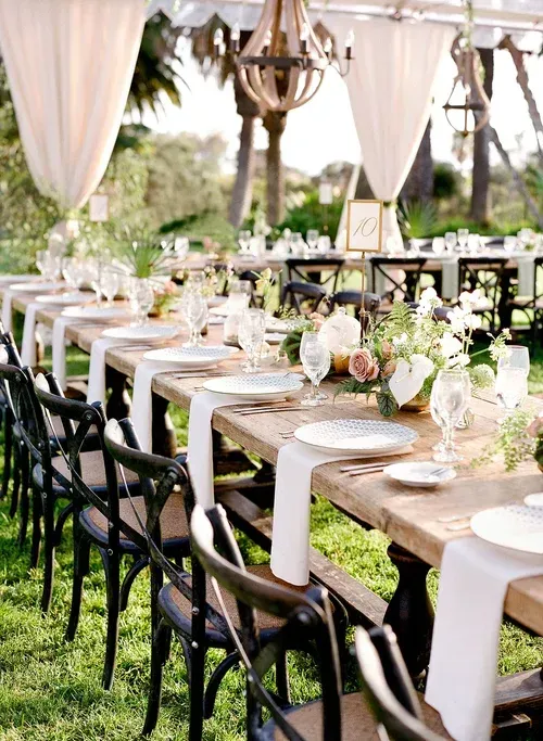 Outdoor wedding reception with long decorated banquet table, black chairs, and white drapes in a garden setting