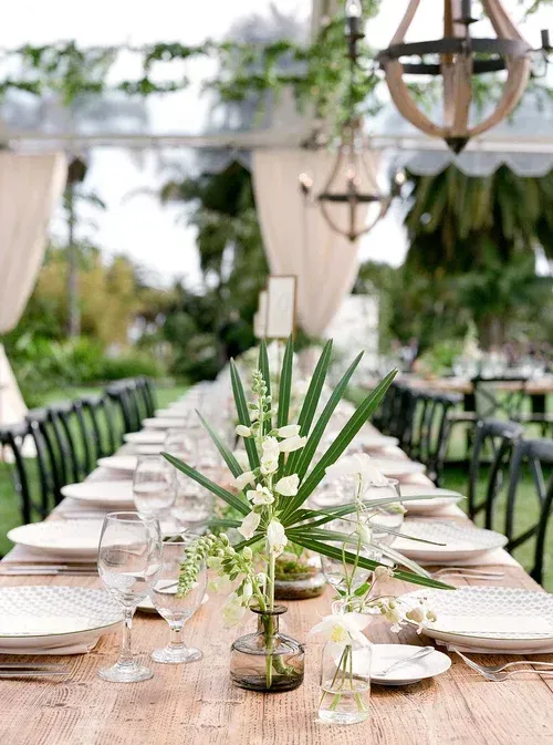 Long outdoor banquet table with white plates, glassware, and greenery under hanging lanterns and draped fabric