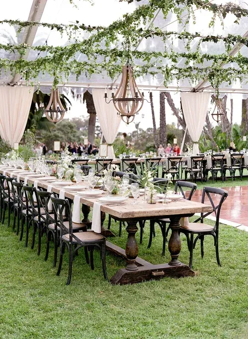 Long banquet tables set under a white tent with greenery and hanging lights on a grassy lawn.