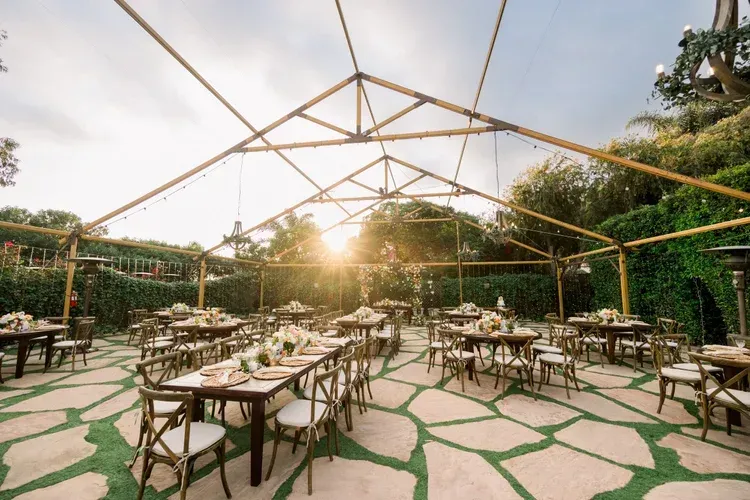 Open-air garden dining setup with tables and chairs under a wooden canopy at sunset