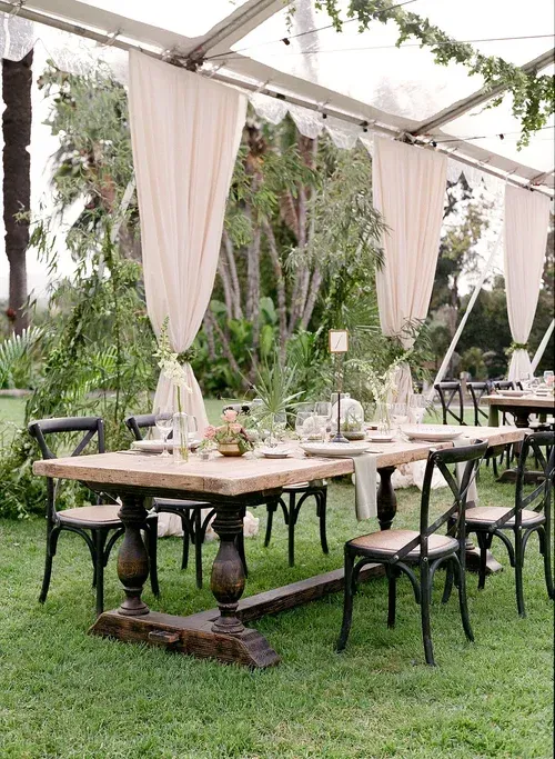 Outdoor rustic reception table under a tent with greenery, draped fabric, and black chairs on grass