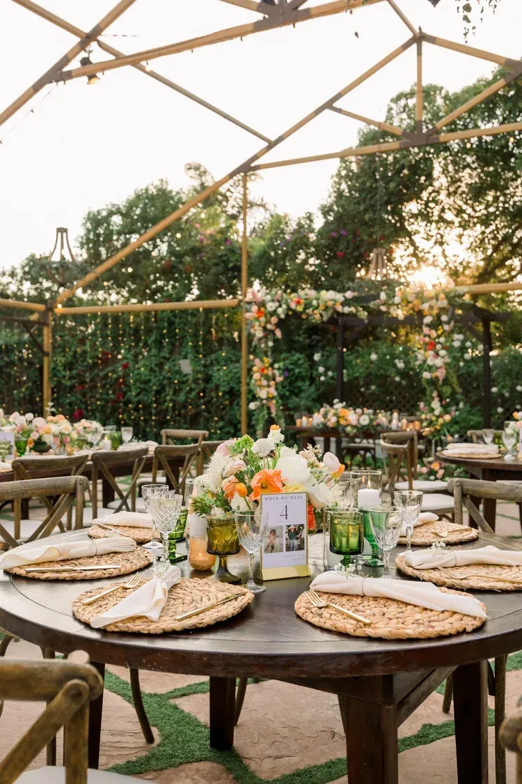 Set outdoor dining table under a clear tent, with flowers, plates, and glassware in a garden setting