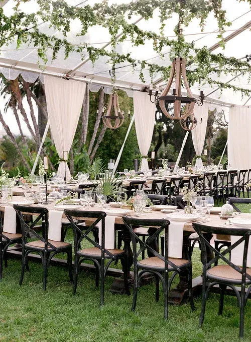 Elegant outdoor dining setup with black chairs, white tablecloths, and draped greenery under a canopy.