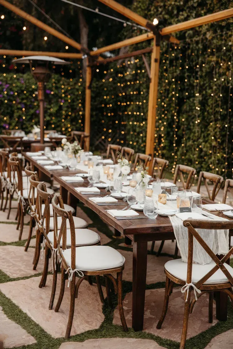 Long banquet table set under a wooden pergola with string lights and greenery backdrop