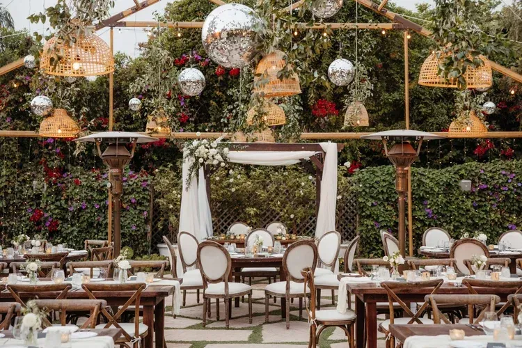 Outdoor wedding reception with white chairs, wooden tables, and hanging lanterns under a floral canopy