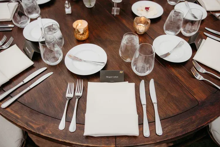 Round wooden dining table set with white plates, folded napkins, silverware, glasses, and candles