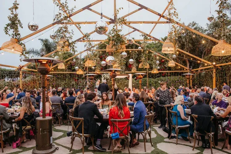 Crowded outdoor market under wooden canopy with hanging plants and people seated at tables