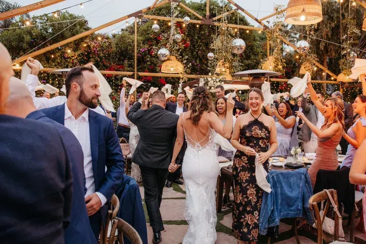 Wedding guests cheering as a bride walks down an outdoor reception aisle with string lights overhead