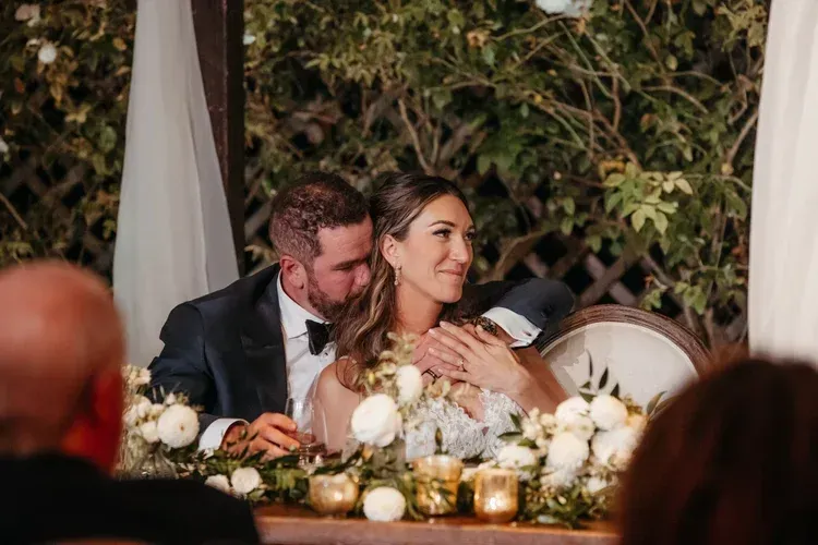 Couple seated at a decorated wedding reception table with white flowers and candles, smiling under warm lights