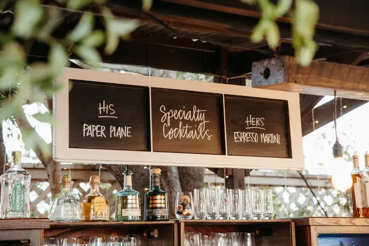 Bar sign with three chalkboard labels above glassware: “His paper plane,” “Specialty cocktails,” and “Hers espresso martini”