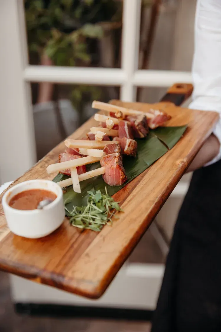 Skewered grilled meat on a wooden board with dipping sauce and greens, served at a table
