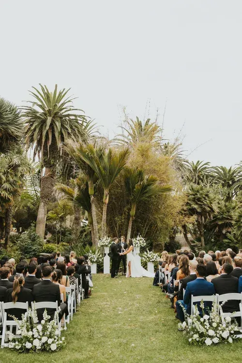 Outdoor wedding ceremony in a palm garden, with guests seated on white chairs along a grass aisle