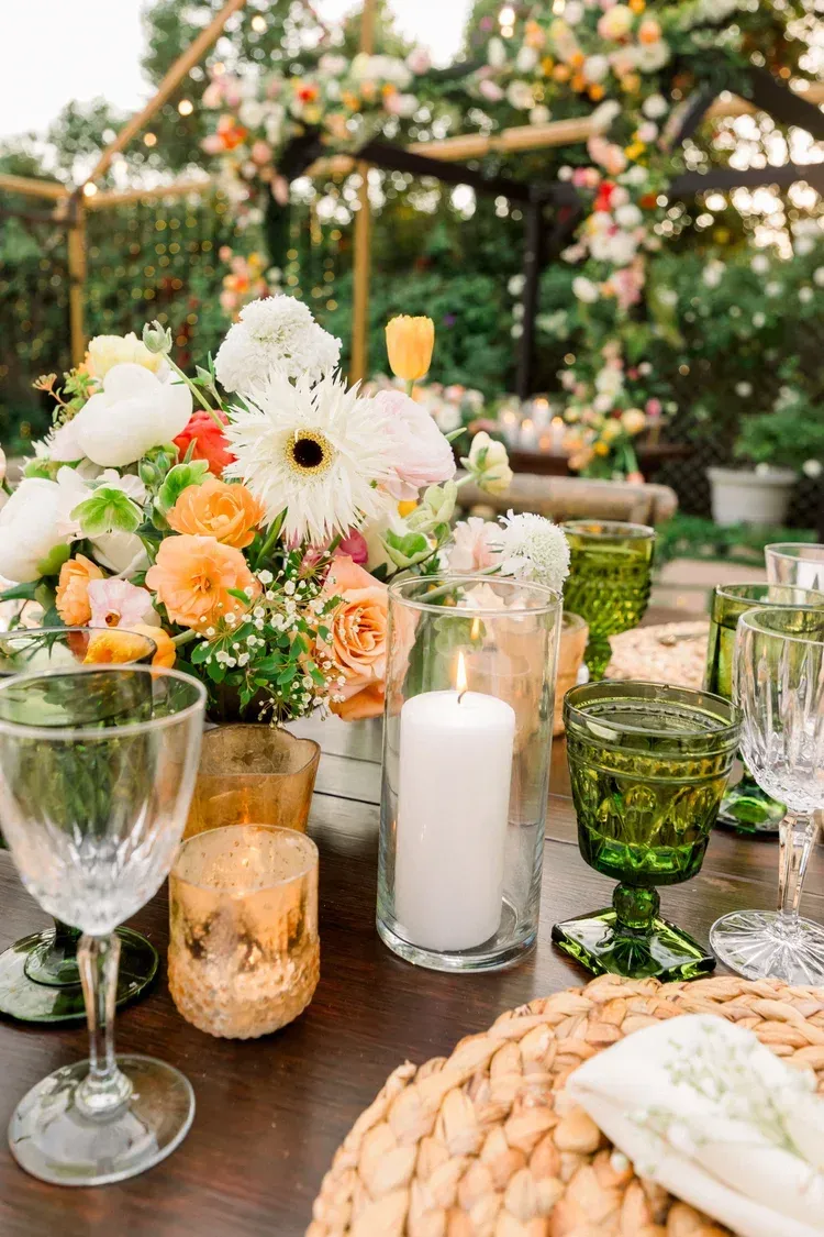 Elegant outdoor table setting with white and peach flowers, glassware, candles, and woven chargers