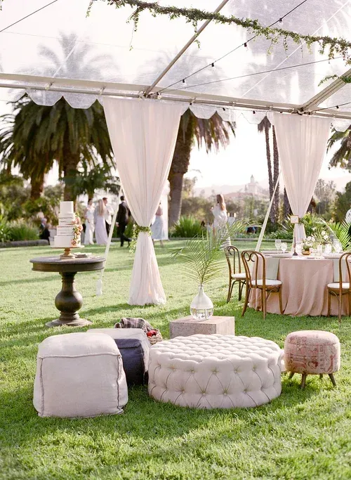 Outdoor wedding lounge with white drapes, tufted ottoman, stools, and tables on a grassy lawn under a canopy