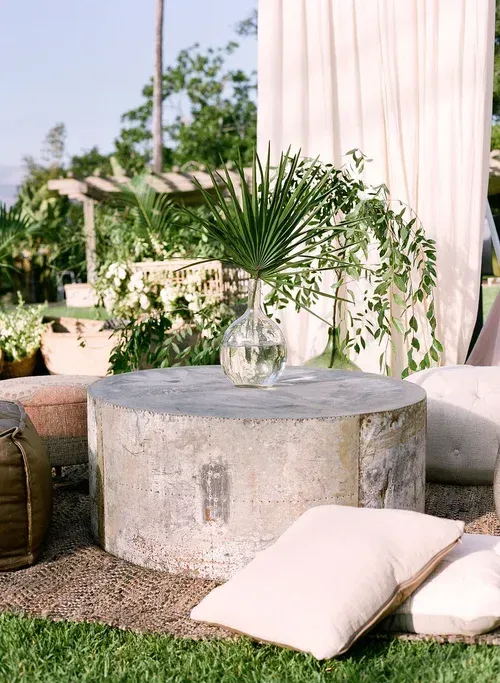 Outdoor lounge with a stone table, glass vase of greenery, and cream cushions under a striped canopy