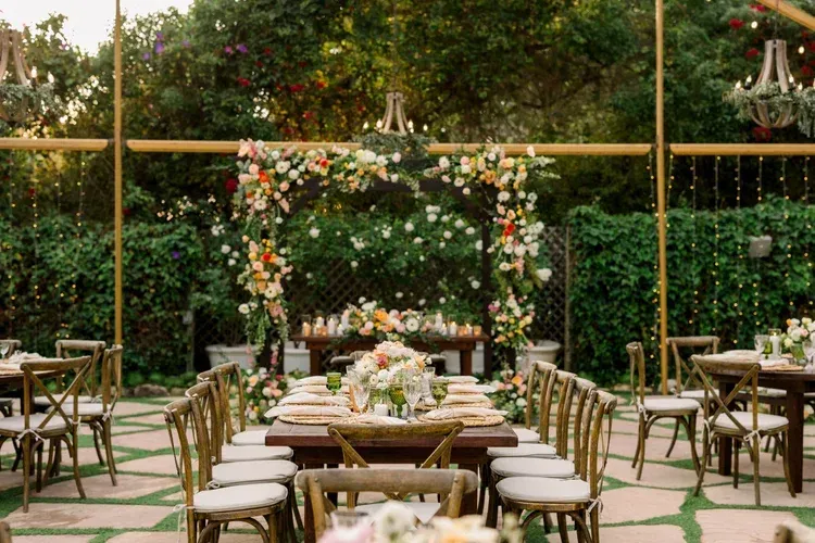 Garden wedding reception setup with floral arch, long tables, and gold chairs under greenery-covered trellis