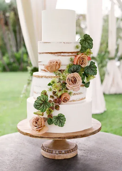 White tiered wedding cake with gold bands and cascading peach roses and greenery on a wooden stand
