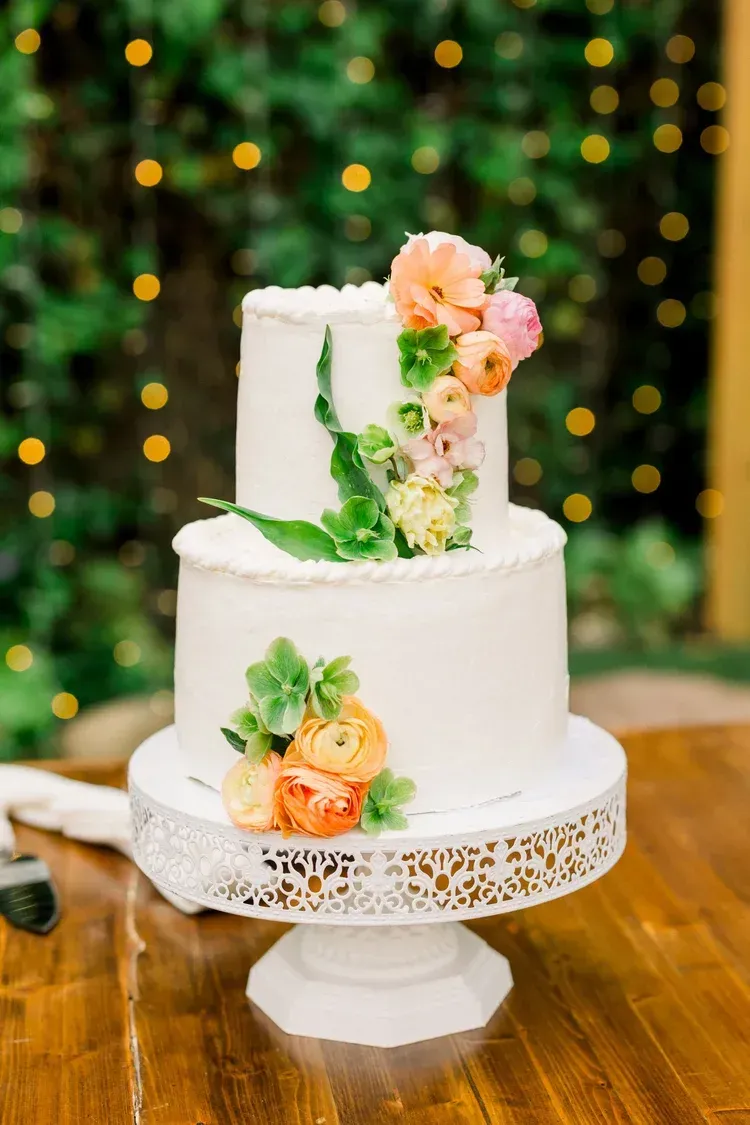 White two-tier wedding cake with peach and pink flowers on a lace stand, against a soft green bokeh backdrop