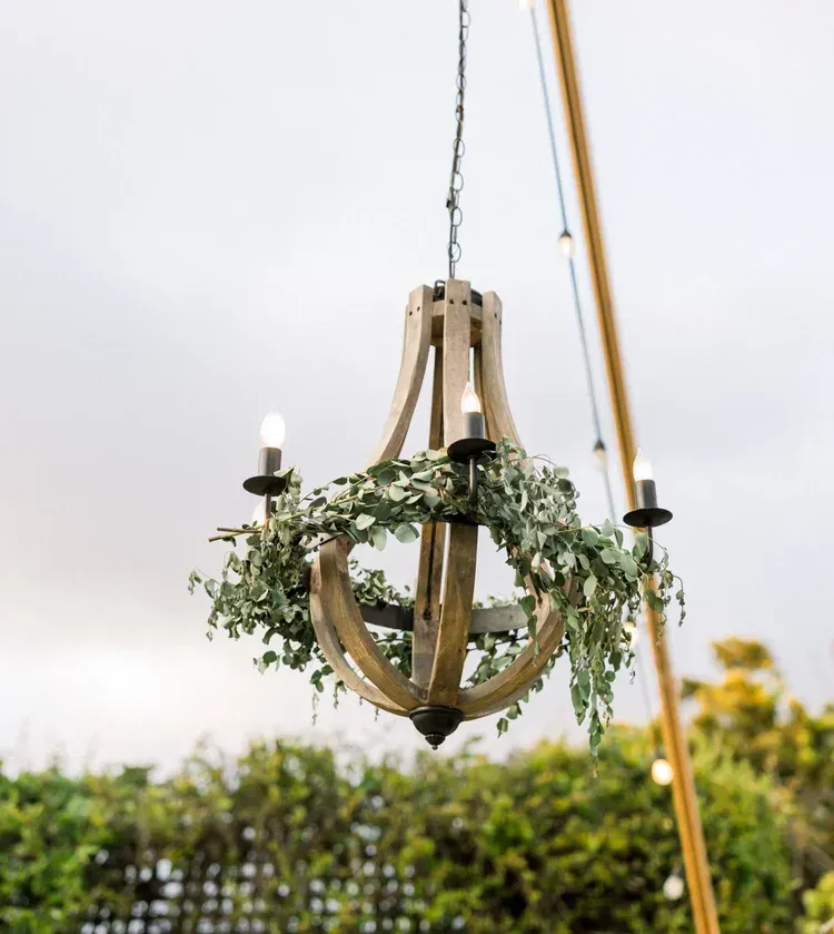 Rustic wooden chandelier with greenery and candles hanging outdoors against a bright sky