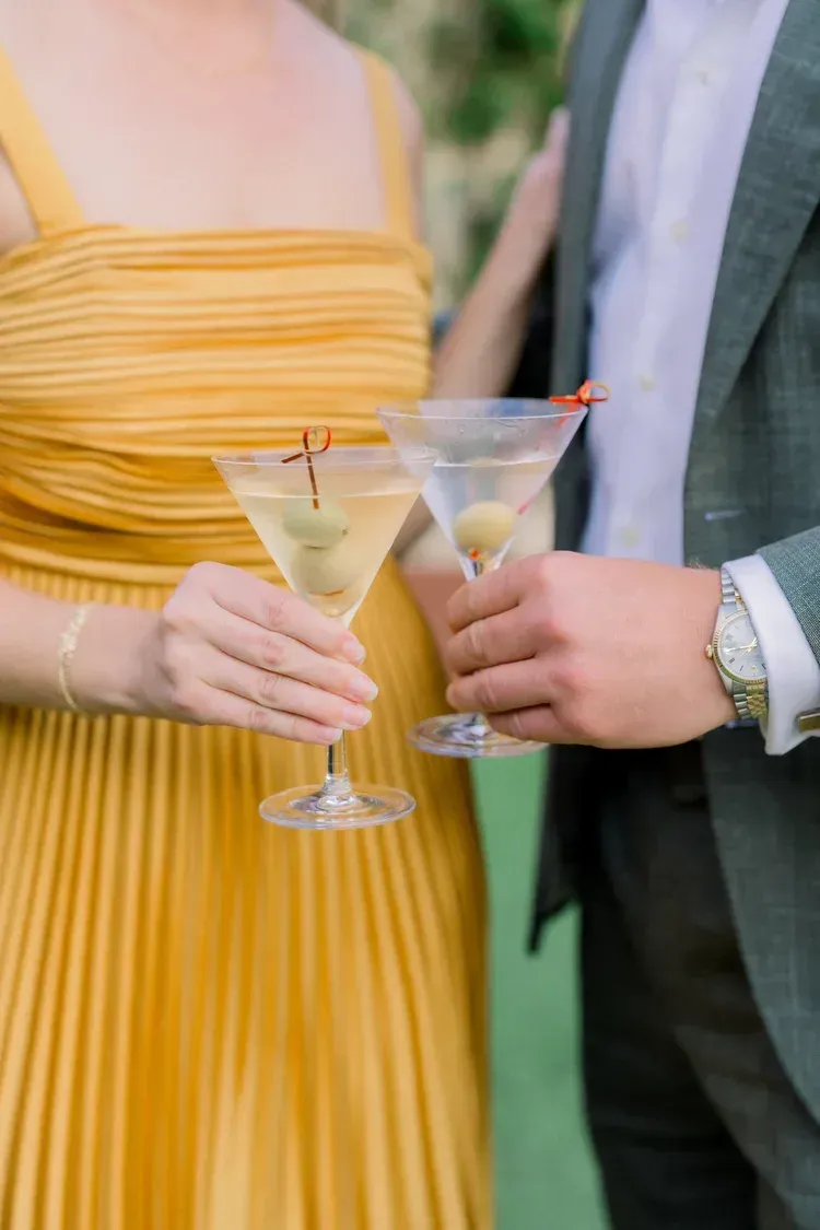 Two people in formal wear hold martini glasses with drinks at a celebration