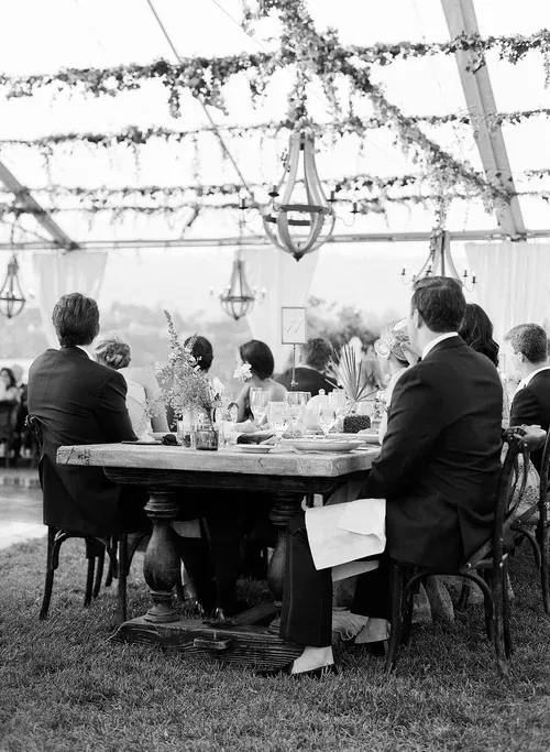 Guests seated at a long table under a decorated outdoor tent, viewed in black and white