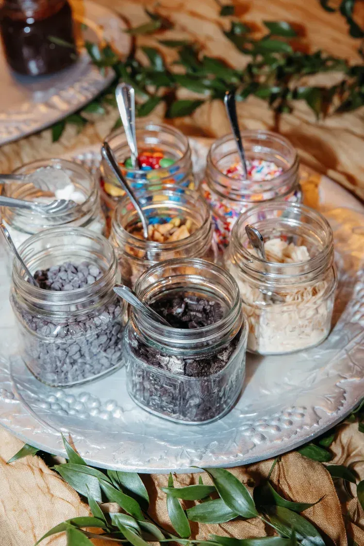 Six small glass jars of condiments on a glass tray, with spoons, on a leafy table setting.