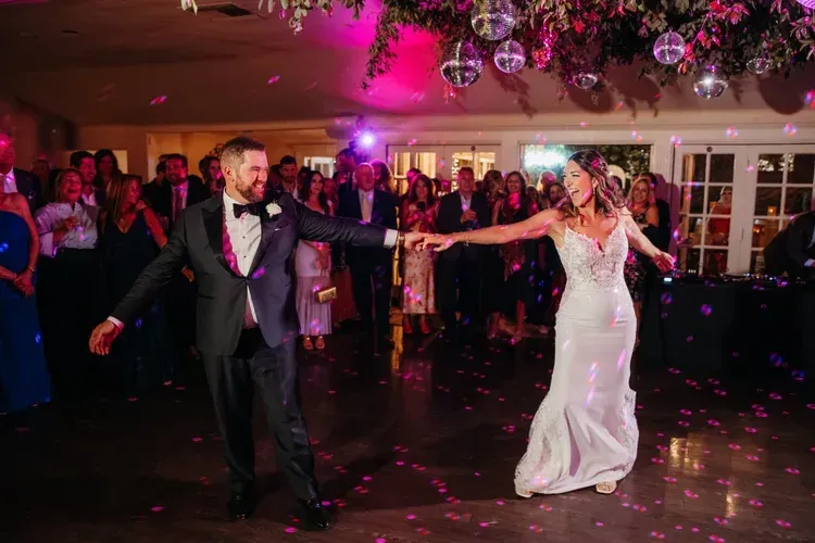 Bride and groom dancing in a decorated reception hall with pink-purple lighting and guests watching