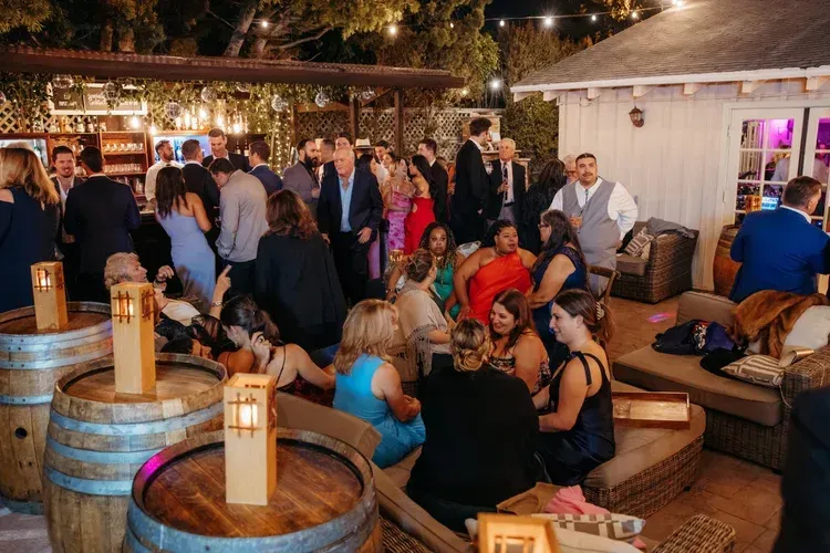 Crowd mingling at an outdoor evening event with wooden barrels, lanterns, and string lights
