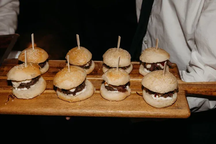 Tray of mini sandwiches on toothpicks, arranged on a wooden board, held by two people