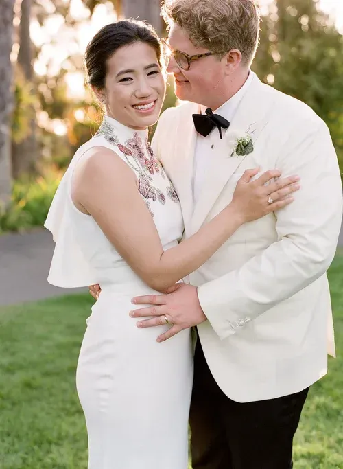 Bride and groom embracing outdoors in a sunlit garden, smiling at the camera.