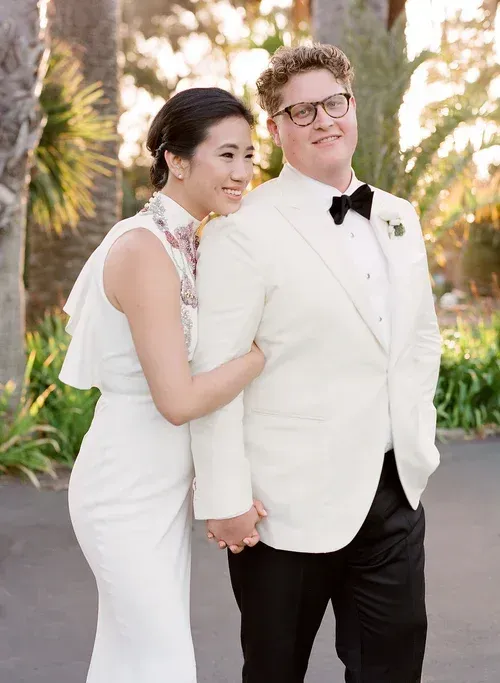 Two people in white formalwear standing outdoors, smiling and holding hands.